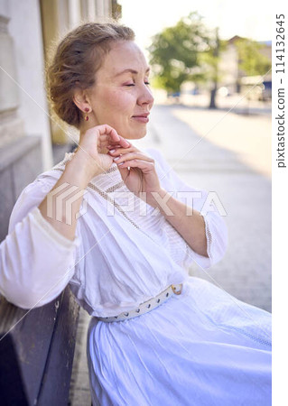 elegant middle age woman in a white vintage dress sitting on a bench in the morning city elegant middle age woman in a white vintage dress sitting on a bench in the morning city 114132645