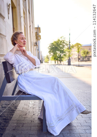 elegant middle age woman in a white vintage dress sitting on a bench in the morning city 114132647