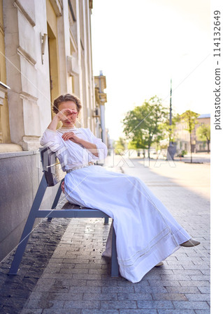 elegant middle age woman in a white vintage dress sitting on a bench in the morning city elegant middle age woman in a white vintage dress sitting on a bench in the morning city 114132649