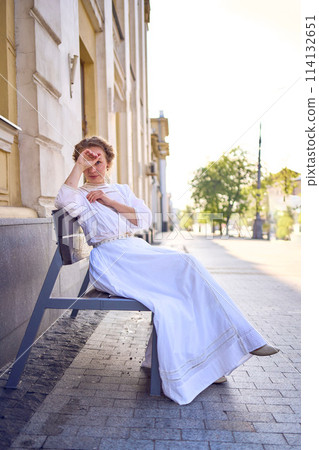 elegant middle age woman in a white vintage dress sitting on a bench in the morning city elegant middle age woman in a white vintage dress sitting on a bench in the morning city 114132651