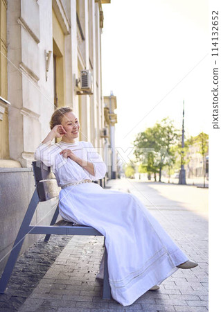elegant middle age woman in a white vintage dress sitting on a bench in the morning city elegant middle age woman in a white vintage dress sitting on a bench in the morning city 114132652
