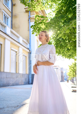 elegant middle age woman in a white vintage dress against the background of historical buildings in the morning light elegant middle age woman in a white vintage dress against the background of historical buildings in the morning light 114132769