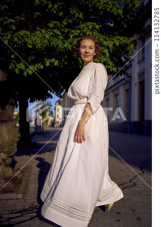 elegant middle age woman in a white vintage dress against the background of historical buildings in the morning light 114132785