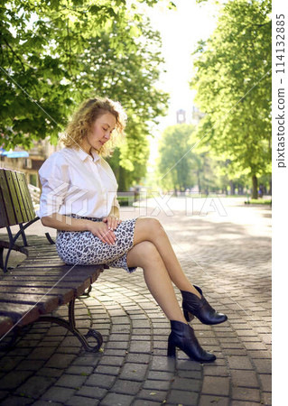 beautiful middle age woman in 70s, 80s style clothes on a bench in a sunlit avenue 114132885