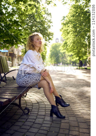 beautiful middle age woman in 70s, 80s style clothes on a bench in a sunlit avenue 114132886