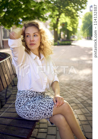 beautiful middle age woman in 70s, 80s style clothes on a bench in a sunlit avenue beautiful middle age woman in 70s, 80s style clothes on a bench in a sunlit avenue 114132887