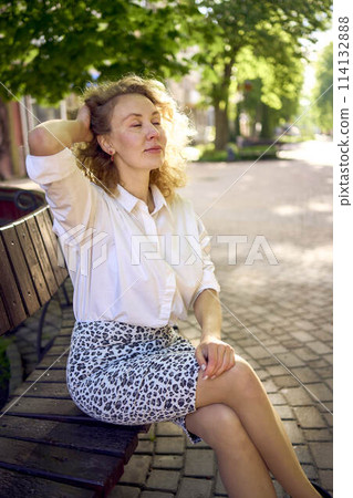 beautiful middle age woman in 70s, 80s style clothes on a bench in a sunlit avenue 114132888