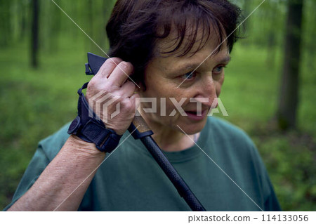 elderly woman engaged in Nordic walking with sticks in spring forest switches music on headphones 114133056