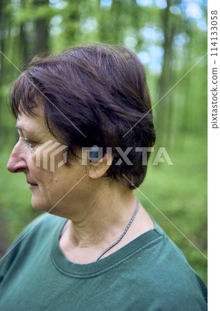 elderly woman engaged in Nordic walking with sticks in spring forest switches music on headphones 114133058