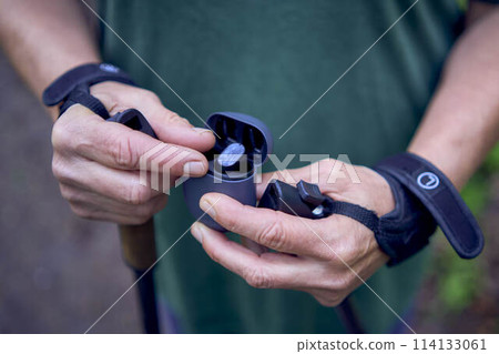 elderly woman engaged in Nordic walking with sticks in spring forest switches music on headphones 114133061