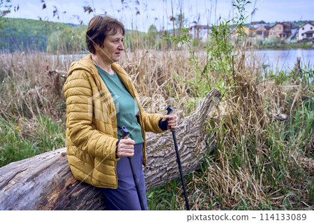 elderly woman rests on the river bank after Nordic walking exercise 114133089