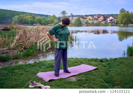 old woman doing exercises in the morning on the river bank, a bicycle and a yogamat next to her 114133121