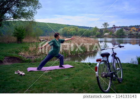 old woman practices yoga in the morning on the river bank, came from home by bicycle old woman practices yoga in the morning on the river bank, came from home by bicycle 114133143