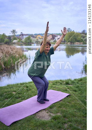 old woman practices yoga in the morning on the river bank, came from home by bicycle 114133146