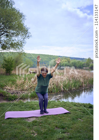old woman practices yoga in the morning on the river bank, came from home by bicycle 114133147
