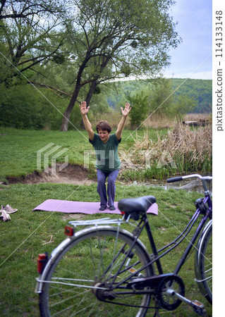 old woman practices yoga in the morning on the river bank, came from home by bicycle 114133148