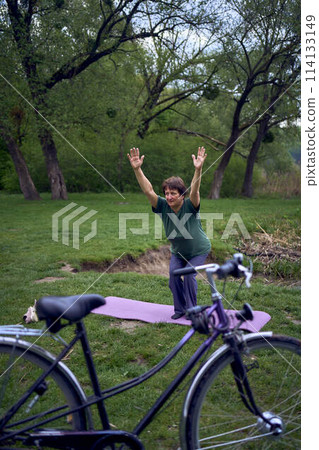 old woman practices yoga in the morning on the river bank, came from home by bicycle old woman practices yoga in the morning on the river bank, came from home by bicycle 114133149