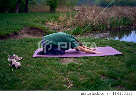 old woman practices yoga in the morning on the river bank, came from home by bicycle 114133155