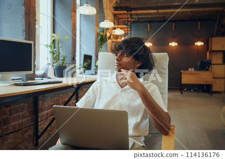 Young man sitting in co-working space, looking thoughtfully away from his laptop, reflecting innovative ideas Young man sitting in co-working space, looking thoughtfully away from his laptop, reflecting innovative ideas 114136176