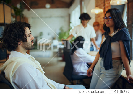 Young people, freelancers occupying a shared workspace, with someone focused on digital screens and others engaged in active discussions. Young people, freelancers occupying a shared workspace, with someone focused on digital screens and others engaged in active discussions. 114136252