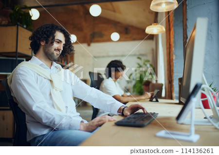 Relaxed co-working space atmosphere. Two young men sitting at table by the window and working on computer. Productive working day Relaxed co-working space atmosphere. Two young men sitting at table by the window and working on computer. Productive working day 114136261