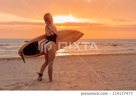 View of young woman surfer girl in swimsuit with surfboard on a sand beach at sunset or sunrise 114137477