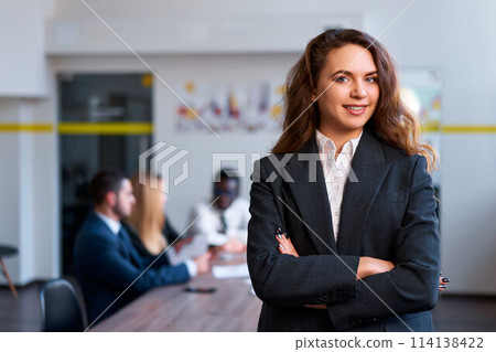 Confident caucasian businesswoman stands in office, arms crossed. Blonde boss oversees team meeting, embodies female leadership. Modern office with diverse staff discussing strategies. 114138422