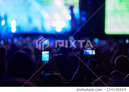 Fans record live music at evening outdoor fest with smartphones, stage lights illuminate excited audience, performers entertain on stage, energetic atmosphere youthful joy, festive celebration. 114138424