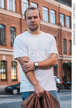 Man in white tshirt holds brown leather jacket outside building Man in white tshirt holds brown leather jacket outside building 114139023