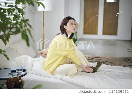 A woman in her twenties trying to put on socks on her bed 114139971