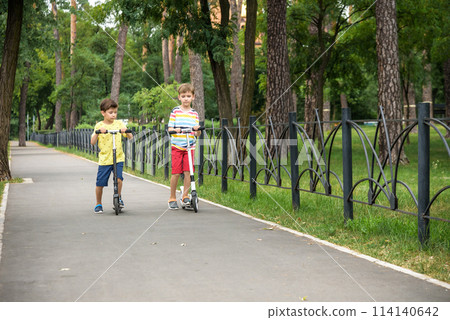 Two attractive European boys brothers, wearing red and white checkered shirts, standing on scooters in the park. They laughing, smiling, hugging and having fun. Active leisure time with kids Two attractive European boys brothers, wearing red and white checkered shirts, standing on scooters in the park. They laughing, smiling, hugging and having fun. Active leisure time with kids 114140642