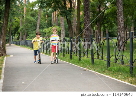 Two attractive European boys brothers, wearing red and white checkered shirts, standing on scooters in the park. They laughing, smiling, hugging and having fun. Active leisure time with kids Two attractive European boys brothers, wearing red and white checkered shirts, standing on scooters in the park. They laughing, smiling, hugging and having fun. Active leisure time with kids 114140643