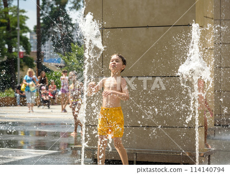Boy having fun in water fountains. Child playing with a city fountain on hot summer day. Happy kids having fun in fountain. Summer weather. Active leisure, lifestyle and vacation 114140794