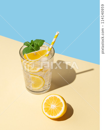 A cold lemonade in a glass with lemon, mint and ice on a yellow and blue background with shadow. A cold lemonade in a glass with lemon, mint and ice on a yellow and blue background with shadow. 114140959