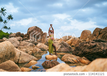 Non-binary black person stands on rock in the middle of still water inside creek. Lgbtq ethnic graceful fashion model poses standing on stone in crystal clear pond at exotic tropical location. 114141121