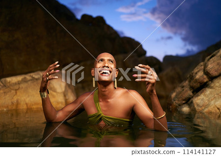 Smiling bipoc lgbtq model poses in water inside natural pool at night. Non-binary person shows jewelry - rings with gems on fingers, brass nose ring, golden earrings, bracelets, stands in pond. Smiling bipoc lgbtq model poses in water inside natural pool at night. Non-binary person shows jewelry - rings with gems on fingers, brass nose ring, golden earrings, bracelets, stands in pond. 114141127