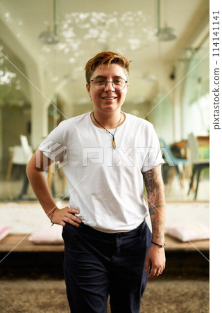 Transgender employee in smart casual attire stands confidently in office environment. Open, modern workspace behind, smiling pro conveys inclusive corporate culture, gender fluidity in business. 114141141