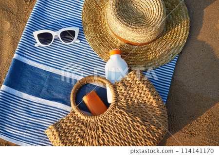Sunbathing accessories on sandy beach, straw hat beside sunglasses, sunblock lotion in bag for skin protection, summer seaside holiday essentials on striped towel, outdoor UV care on sunny day. 114141170