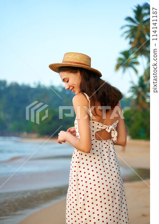 Woman in polka dot dress enjoys sunrise on sandy beach. Boho straw hat, vintage summer style. Solo traveler cherishes ocean view, tropical vacation vibes. Seaside leisure, peaceful morning by sea. 114141187