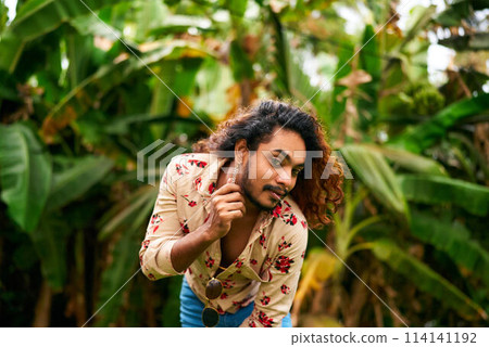 Confident gay man with ethnic background poses in rich tropical setting, smiling and touching earring. Expressive male in printed shirt embodies pride and fashion, surrounded by plants. 114141192