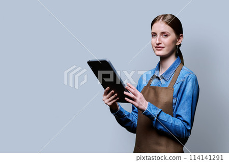 Young female worker in apron holding digital tablet on gray studio background 114141291