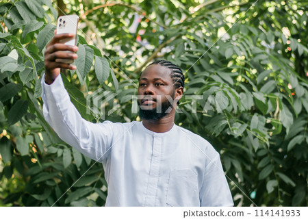 Happy young African American man in dashiki ethnic clothes taking selfie on green leaves outdoor background. Millennial generation student and youth. Happy young African American man in dashiki ethnic clothes taking selfie on green leaves outdoor background. Millennial generation student and youth. 114141933