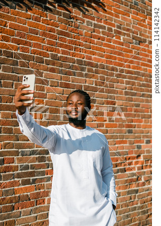 Happy young African American man in dashiki ethnic clothes taking selfie on brick wall background. Millennial generation student and youth. 114142342