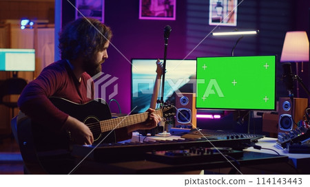 Young artist playing his guitar and practicing new song with online tutorial, looks at computer with greenscreen display. Musician learning new acoustic instrument accords, home studio. Camera A. 114143443