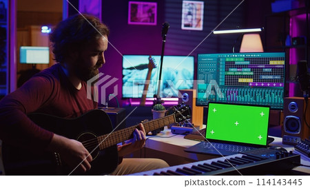 Musician beginner learning accords to play acoustic guitar by watching tutorials and online lessons, laptop shows greenscreen chromakey. Artist practicing guitar skills in home studio. Camera B. Musician beginner learning accords to play acoustic guitar by watching tutorials and online lessons, laptop shows greenscreen chromakey. Artist practicing guitar skills in home studio. Camera B. 114143445