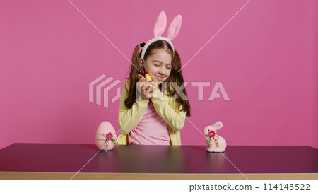 Joyful little girl playing with festive easter decorations in studio, creating arrangements with a chick, rabbit and egg. Smiling cute toddler with bunny ears showing colorful ornaments. Camera B. Joyful little girl playing with festive easter decorations in studio, creating arrangements with a chick, rabbit and egg. Smiling cute toddler with bunny ears showing colorful ornaments. Camera B. 114143522