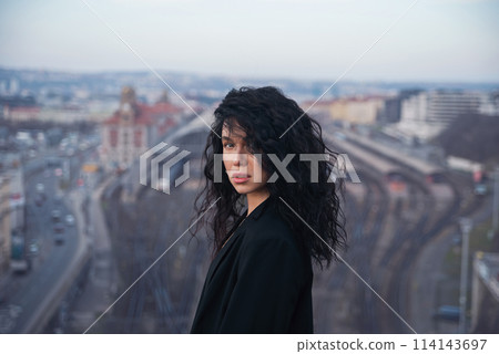 Attractive young brunette woman in a black jacket stands on a rooftop against a city space background. 114143697