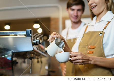 Professional Asian female barista pouring a hot milk over the espresso in coffee cup. Happy female barista making a cup of latte coffee in a small coffee shop. Small business concept. Professional Asian female barista pouring a hot milk over the espresso in coffee cup. Happy female barista making a cup of latte coffee in a small coffee shop. Small business concept. 114146084