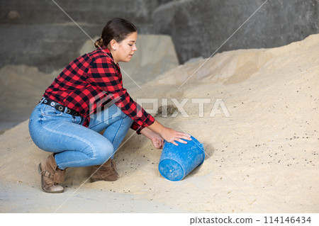 Happy young woman farmer in plaid shirt squatting with handful of cornmeal in her hands in storage 114146434