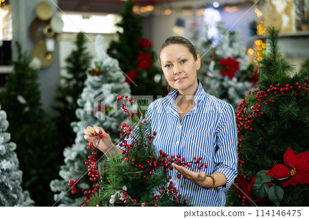 Positive middle-aged female choosing an artificial Christmas tree decorated with viburnum at the December 114146475
